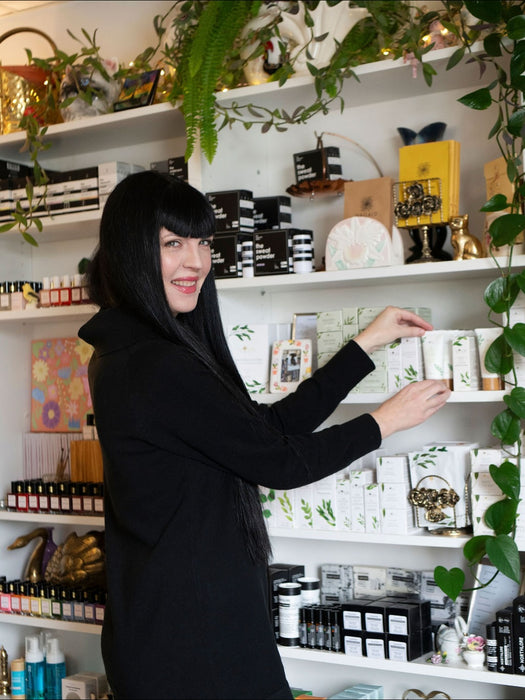Jennilee Cardinal-Schultz Saskatoon Bridal Makeup Artist. Woman in a black coat standing in a store with shelves filled with products and plants.